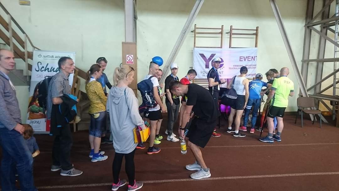 Grupa osób w hali sportowej, prawdopodobnie po biegu, niektórzy piją wodę, w tle widoczny baner z napisem 'EVENT' i logo firmy.