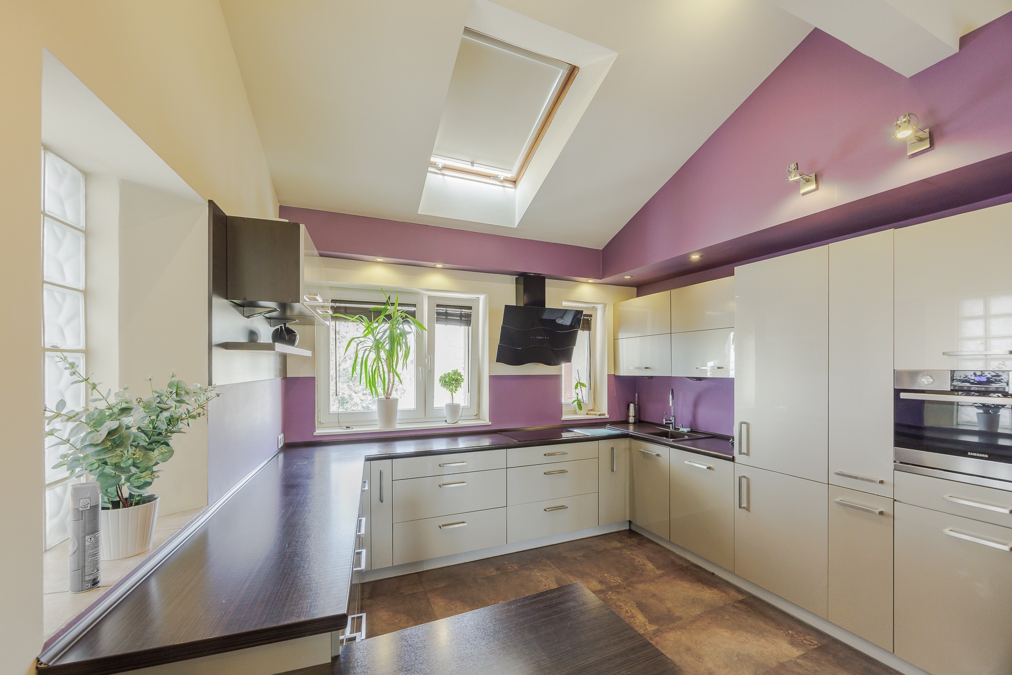 Nowoczesna kuchnia z białymi szafkami, ciemnym blatem i fioletowymi akcentami na ścianach, oświetlona światłem wpadającym przez okno dachowe.
