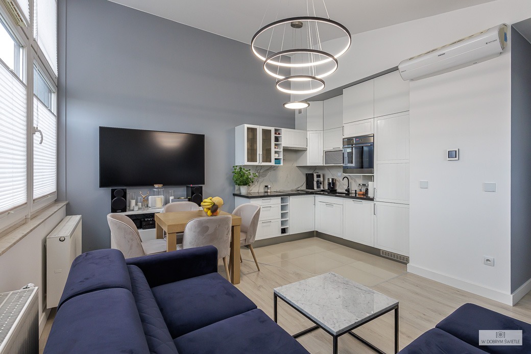 Nowoczesne wnętrze mieszkania z otwartą kuchnią, białymi meblami kuchennymi i wyspą, drewnianym stołem jadalnym, granatową sofą i okrągłą lampą sufitową z trzema pierścieniami.