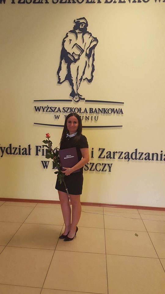 Kobieta w czarnej sukience trzymająca czerwoną różę i fioletową pracę magisterską na tle logo Wyższej Szkoły Bankowej w Gdańsku, Wydział Finansów i Zarządzania.