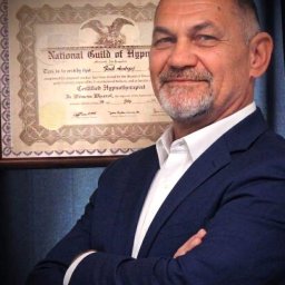 Hipnoza Regresyjna - Uśmiechnięty mężczyzna w granatowym garniturze, z siwą brodą, ze skrzyżowanymi ramionami, na tle oprawionego certyfikatu 'Certified Hypnotherapist' z National Guild of Hypnotists.