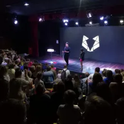 Iluzjonista w czarnym stroju i cylindrze na scenie z młodą asystentką podczas występu dla publiczności w teatrze, oświetlenie sceniczne i logo na ekranie w tle.