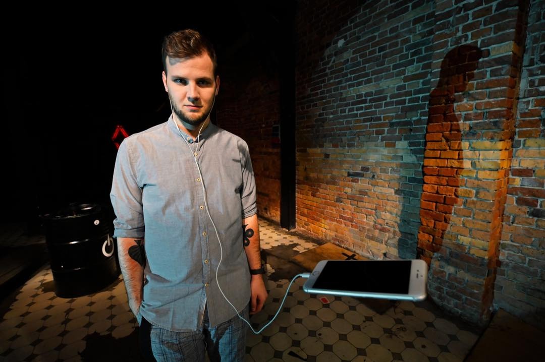 Młody mężczyzna w szarej koszuli z podwiniętymi rękawami, słuchawkami w uszach, stojący na tle ceglanej ściany i podłogi z wzorem, z unoszącym się w powietrzu smartfonem podłączonym kablem. W tle...
