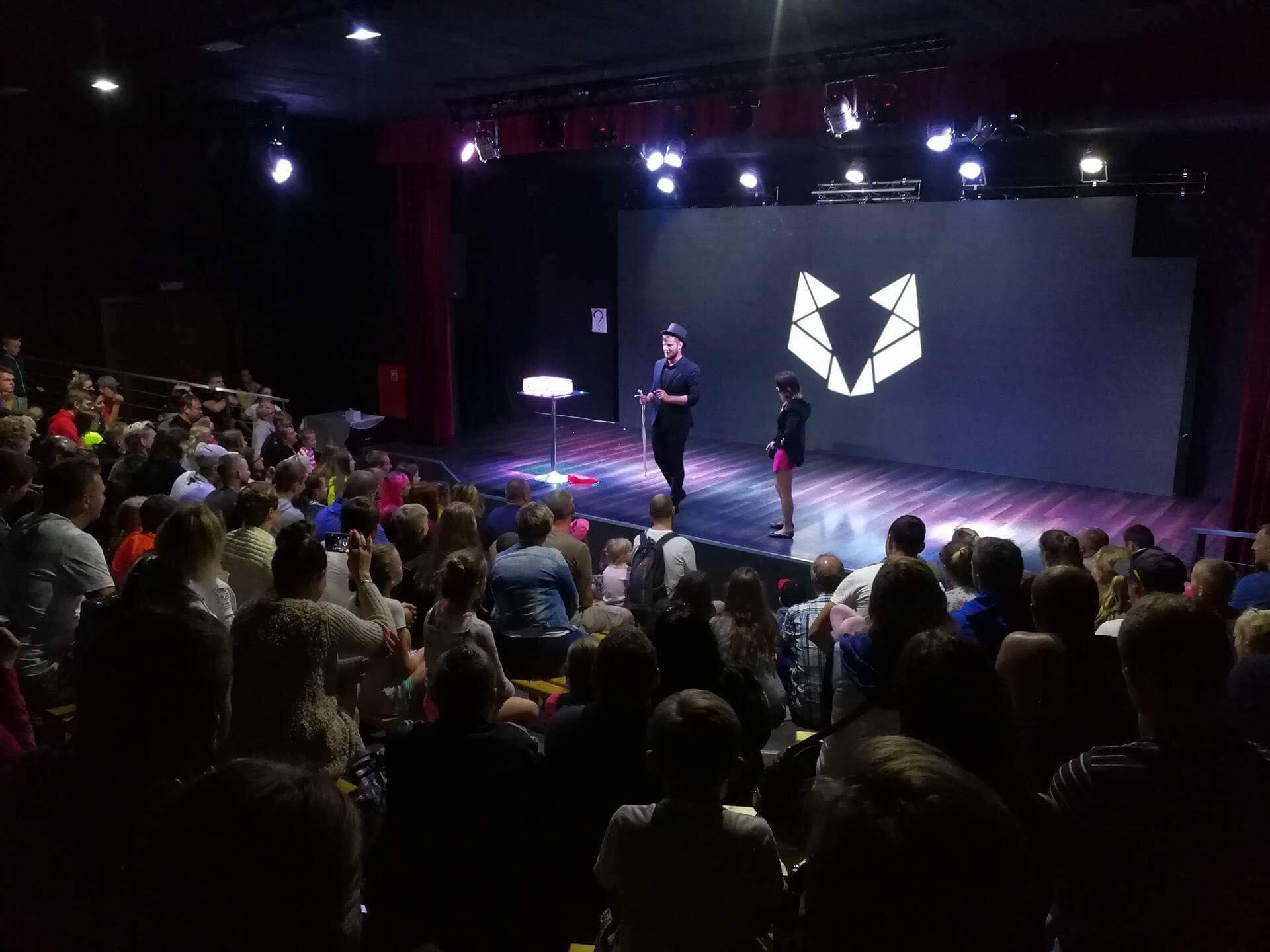 Iluzjonista w czarnym stroju i cylindrze na scenie z młodą asystentką podczas występu dla publiczności w teatrze, oświetlenie sceniczne i logo na ekranie w tle.