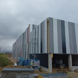 Montaż elewacji hali przemysłowej z wykorzystaniem podnośników koszowych; widoczne panele elewacyjne w odcieniach szarości, biały i czerwony, drabina serwisowa na ścianie, teren budowy w trakcie...