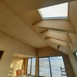 EUROTYNK - Jasne wnętrze po nałożeniu tynku, z oknami dachowymi i dużymi przeszkleniami. Widoczne drabiny i sprzęt tynkarski. Świeżo położony tynk o jednolitej fakturze.