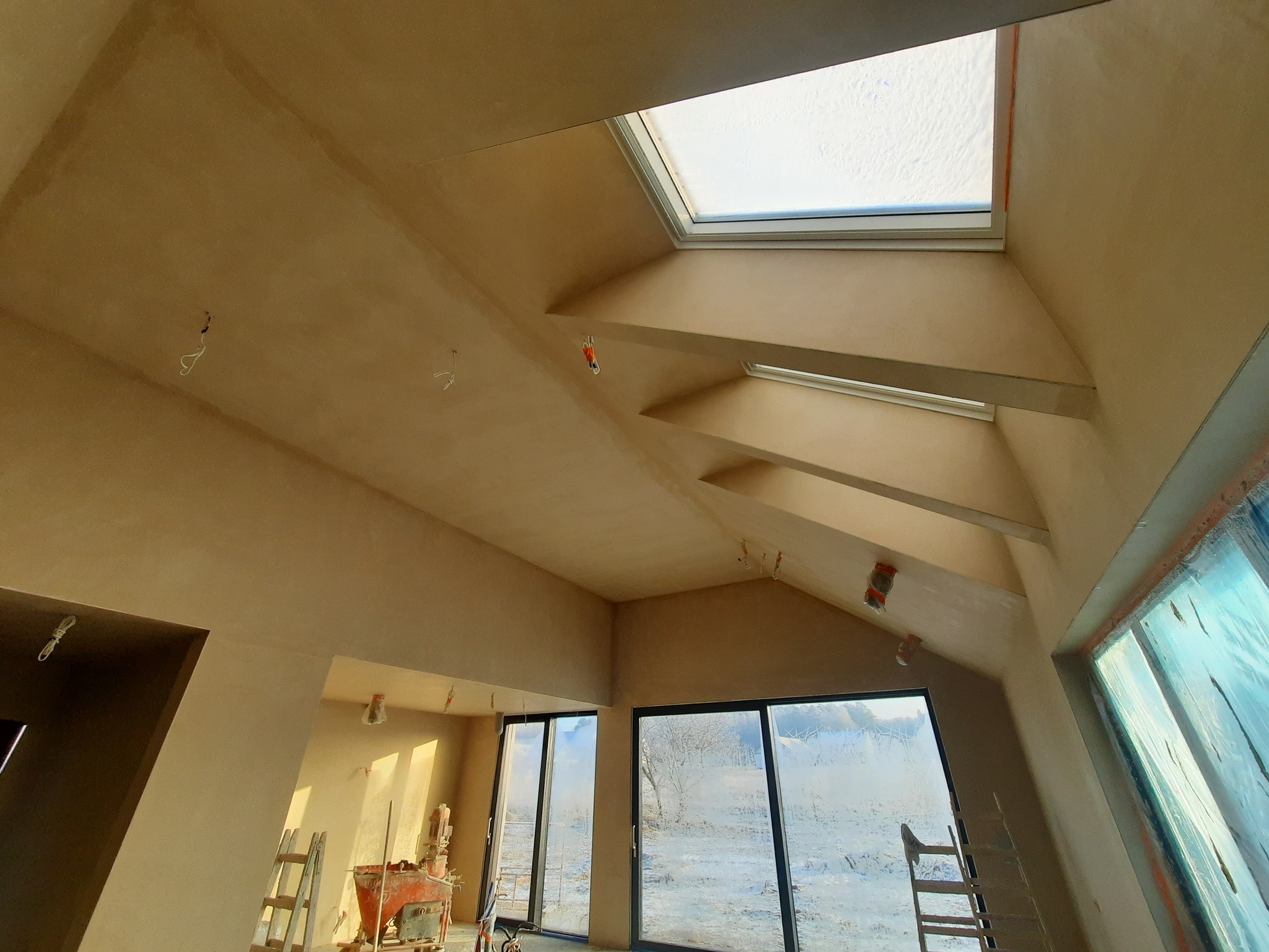 Jasne wnętrze po nałożeniu tynku, z oknami dachowymi i dużymi przeszkleniami. Widoczne drabiny i sprzęt tynkarski. Świeżo położony tynk o jednolitej fakturze.
