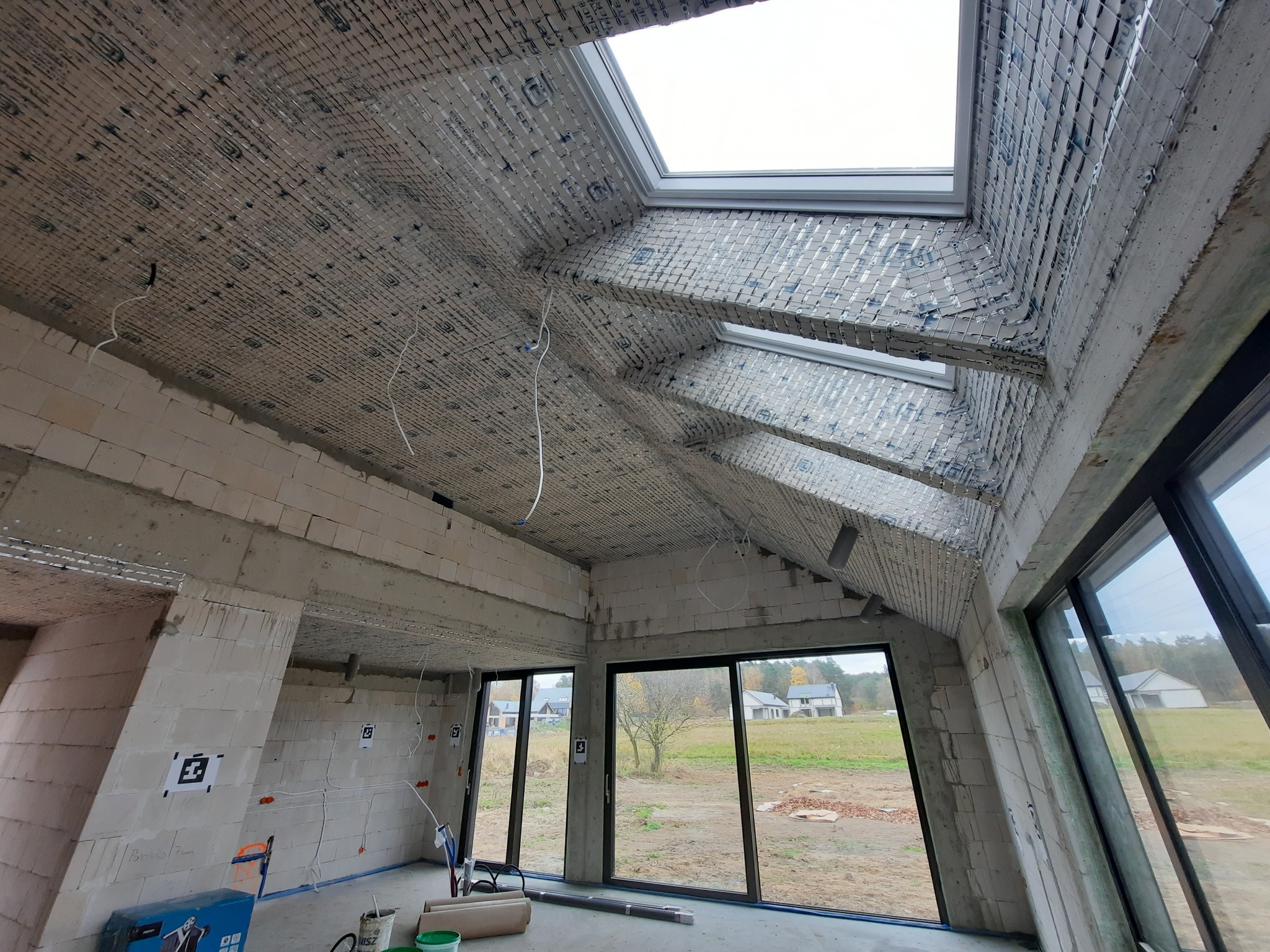 Surowy stan wnętrza z oknami dachowymi i dużymi przeszkleniami. Widoczne bloczki, instalacje i siatka na suficie, przygotowane do dalszych prac wykończeniowych.