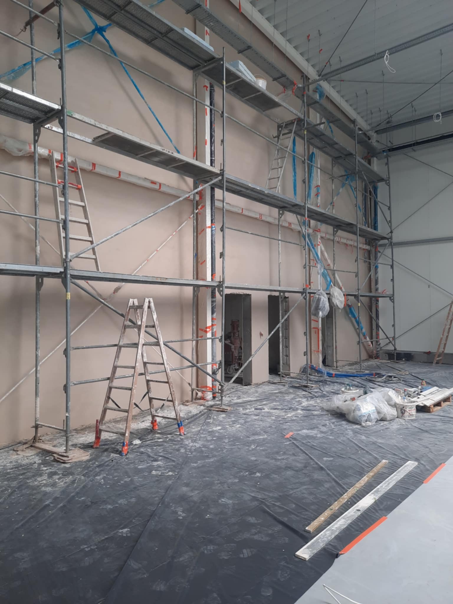 Ściana po nałożeniu tynku w hali przemysłowej, widoczne rusztowania i drabiny. Podłoga zabezpieczona folią. Świeży tynk ma jasny, beżowy odcień.