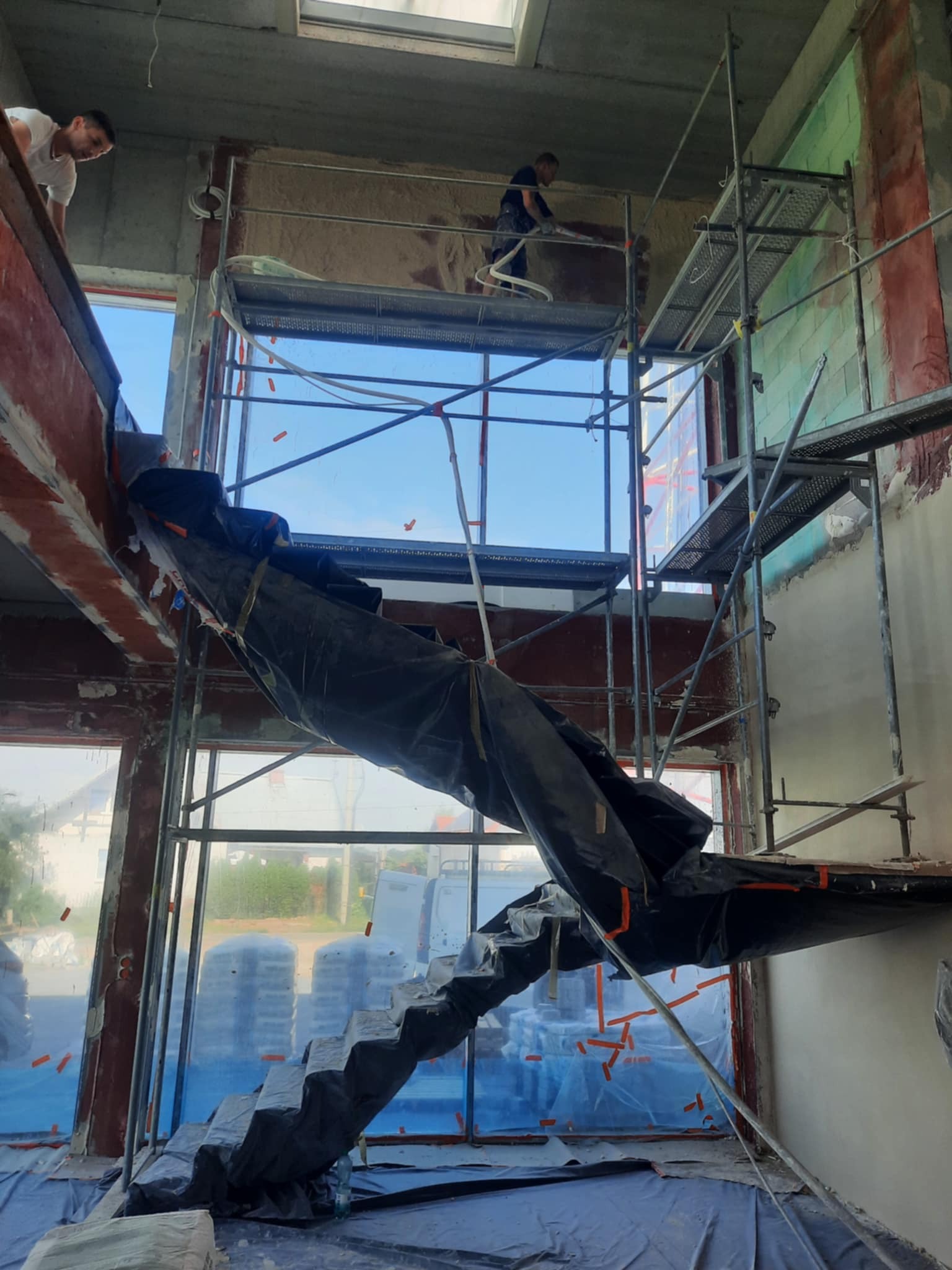 Prace tynkarskie w wysokim pomieszczeniu: robotnik na rusztowaniu aplikuje tynk maszynowy, zabezpieczone schody folią, widoczne okna i worki z materiałem.