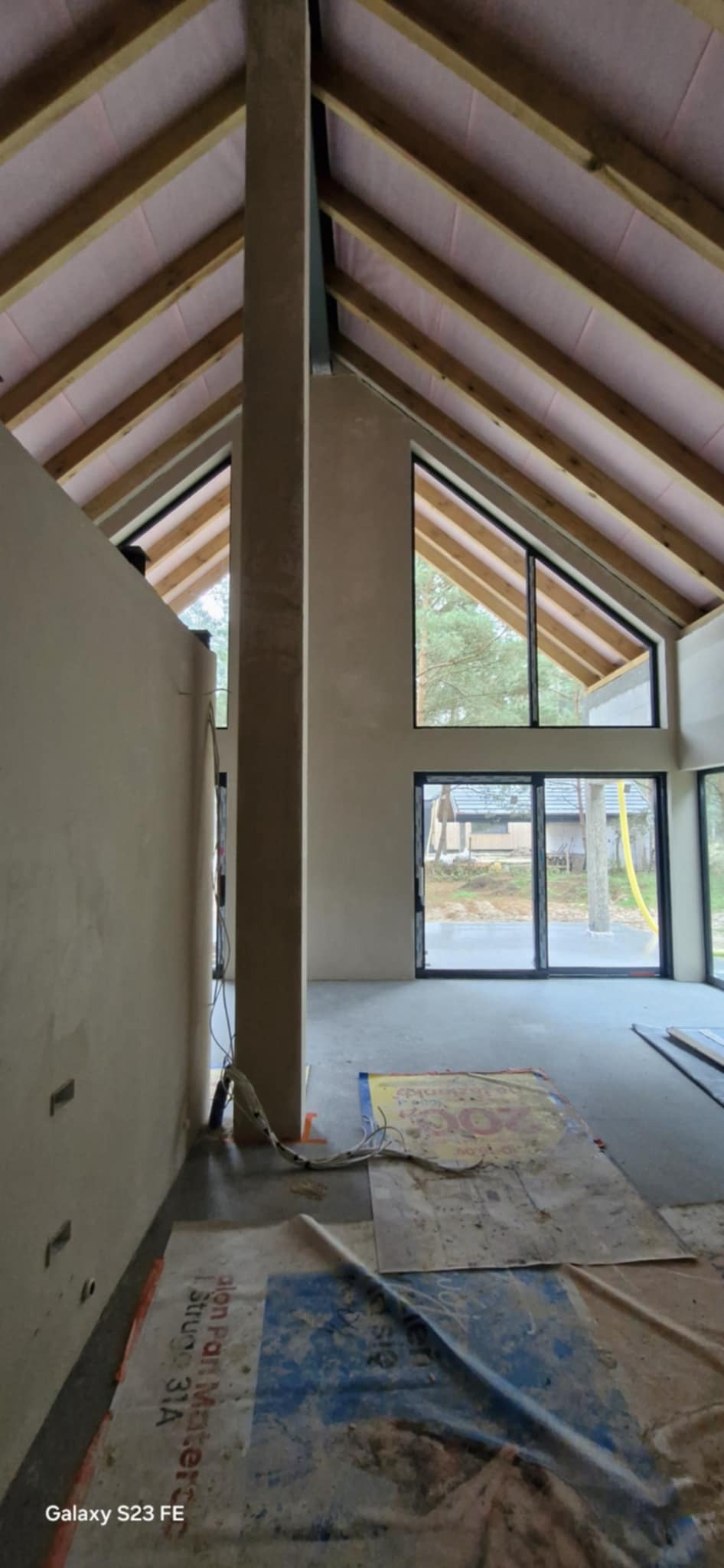 Surowe wnętrze domu w trakcie prac wykończeniowych. Widoczne ściany po nałożeniu tynku, duże okna z widokiem na zewnątrz i drewniana konstrukcja dachu.