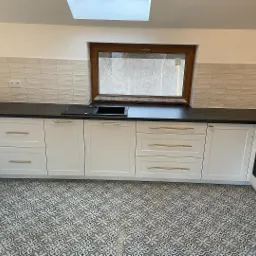 Nowoczesna kuchnia z białymi szafkami, czarnym blatem i wzorzystą podłogą. Okno z brązową ramą nad zlewem. Ściana wyłożona płytkami imitującymi cegłę.