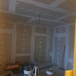 Pokój w trakcie wykańczania: ściany i sufit pokryte płytami gipsowo-kartonowymi, widoczne szpachlowane łączenia, narzędzia i materiały budowlane w pomieszczeniu.