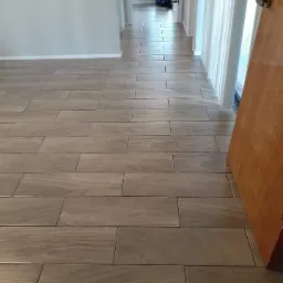 Ułożona podłoga z podłużnych, brązowych płytek imitujących drewno, widok na korytarz z białymi ścianami i drzwiami po prawej stronie.