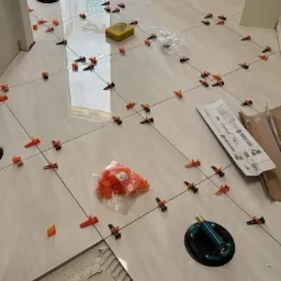 Układanie jasnych płytek podłogowych z użyciem systemu poziomowania płytek, widoczne przyssawki do przenoszenia płytek, paczka z klinami i gąbka do czyszczenia fug.