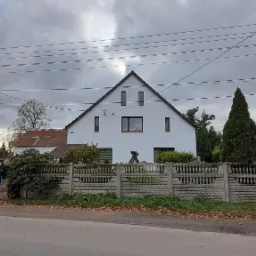 Biały, jednopiętrowy dom z szarym betonowym ogrodzeniem w pochmurny dzień, widoczne linie energetyczne i drzewa iglaste