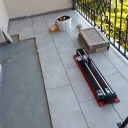 Balkon w trakcie kładzenia szarych płytek, widoczna przecinarka do płytek, wiadro z akcesoriami i paczka płytek.