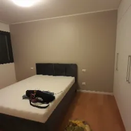 Wnętrze sypialni po malowaniu ścian na szaro, z zamontowanym oknem w czarnej ramie, nowym ł&oacute;żkiem i białą szafą wnękową.