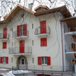 Odnowiony budynek wielorodzinny z kremową elewacją, czerwonymi okiennicami i kutymi balkonami, widok z poziomu gruntu, gałęzie drzew na pierwszym planie.