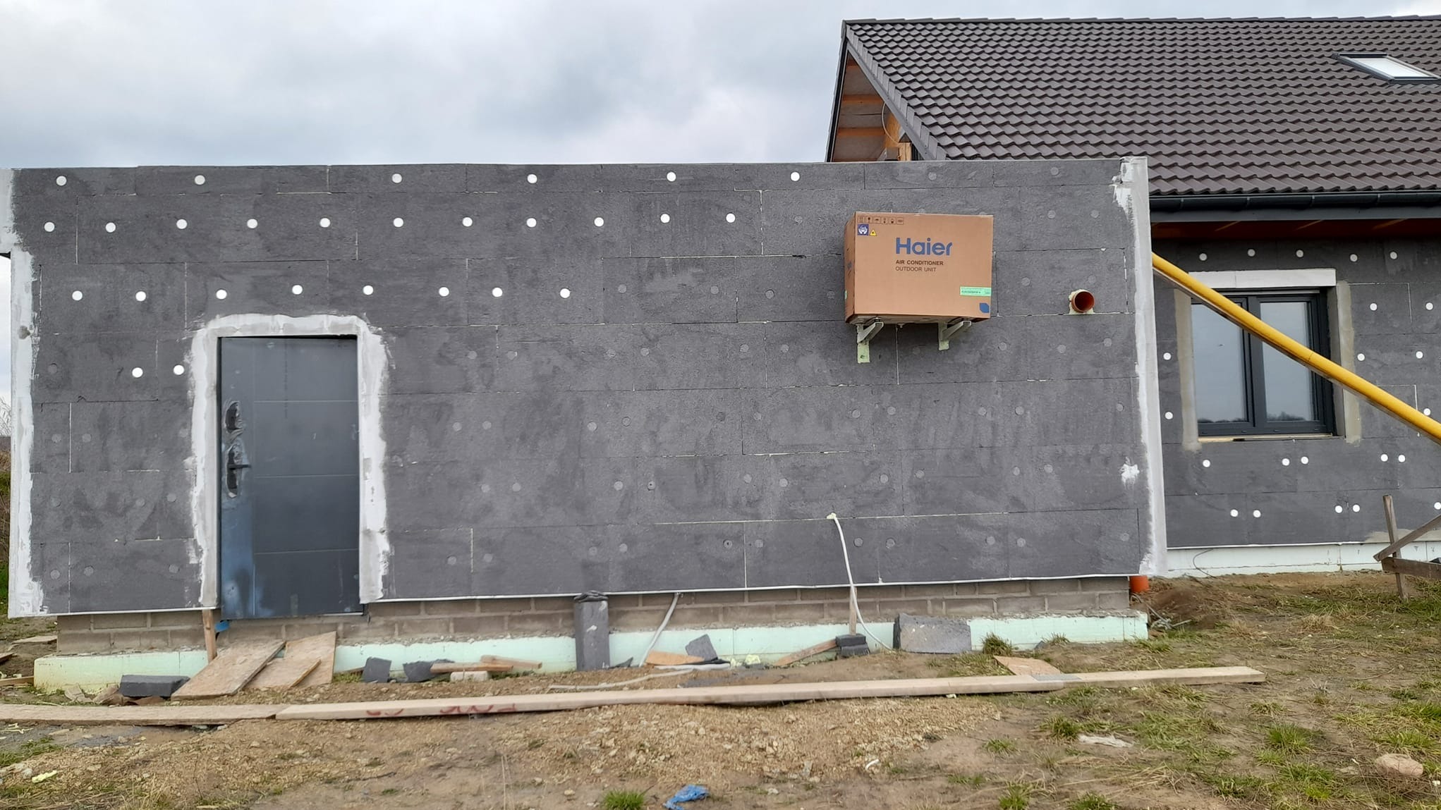 Ściana budynku w trakcie ocieplania styropianem grafitowym, widoczna zamontowana jednostka zewnętrzna klimatyzacji w kartonie, częściowo ocieplone okno oraz czarne drzwi wejściowe.