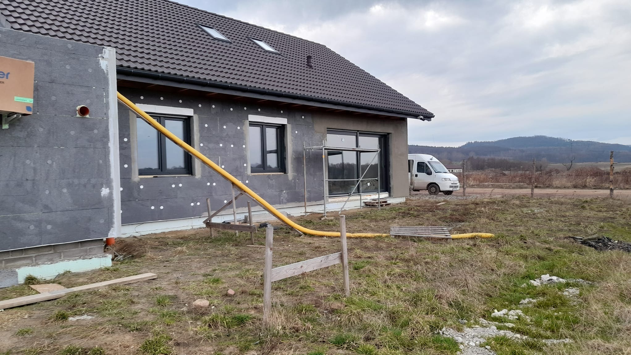 Dom jednorodzinny w trakcie ocieplania, widoczne szare płyty izolacyjne na ścianach, żółty wąż wentylacyjny oraz rusztowanie przy oknach, w tle wzgórza i biały bus.