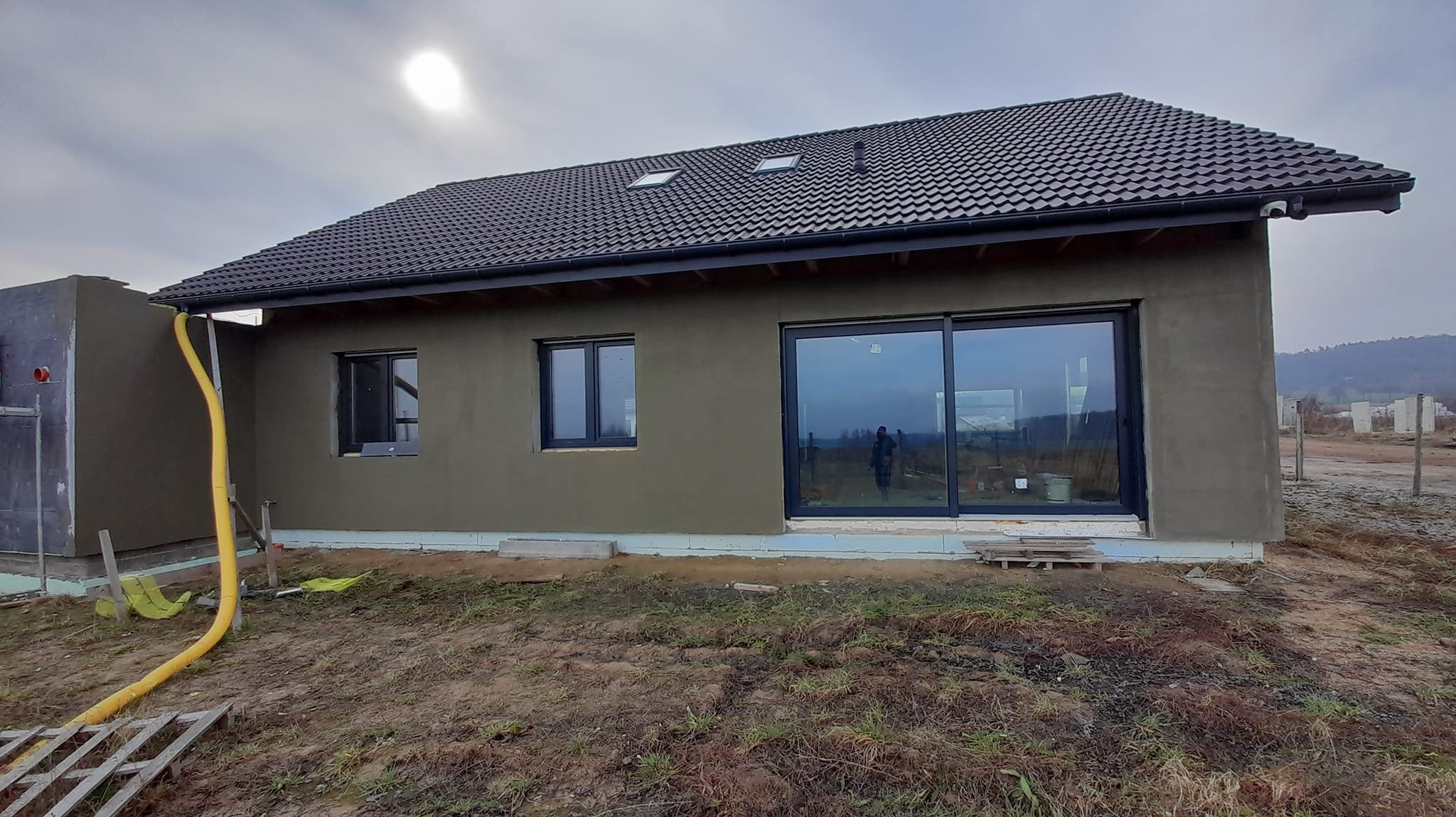 Dom jednorodzinny w trakcie ocieplania, widoczna elewacja pokryta szarą warstwą izolacyjną, czarne okna, dach z ciemnej dachówki i żółty wąż doprowadzający materiał izolacyjny.