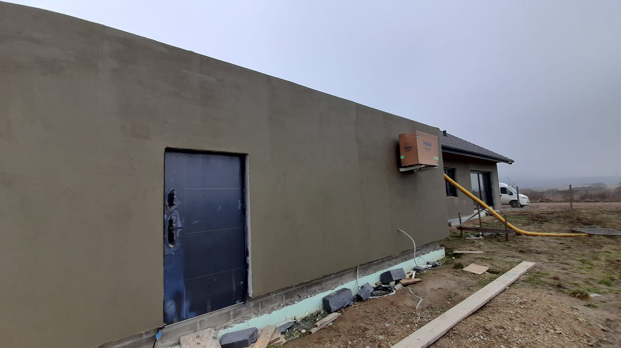 Świeżo otynkowana ściana domu z widocznym otworem drzwiowym zabezpieczonym folią, z zamontowanym na zewnątrz klimatyzatorem w kartonie i żółtym wężem doprowadzającym powietrze, w tle zaparkowany...