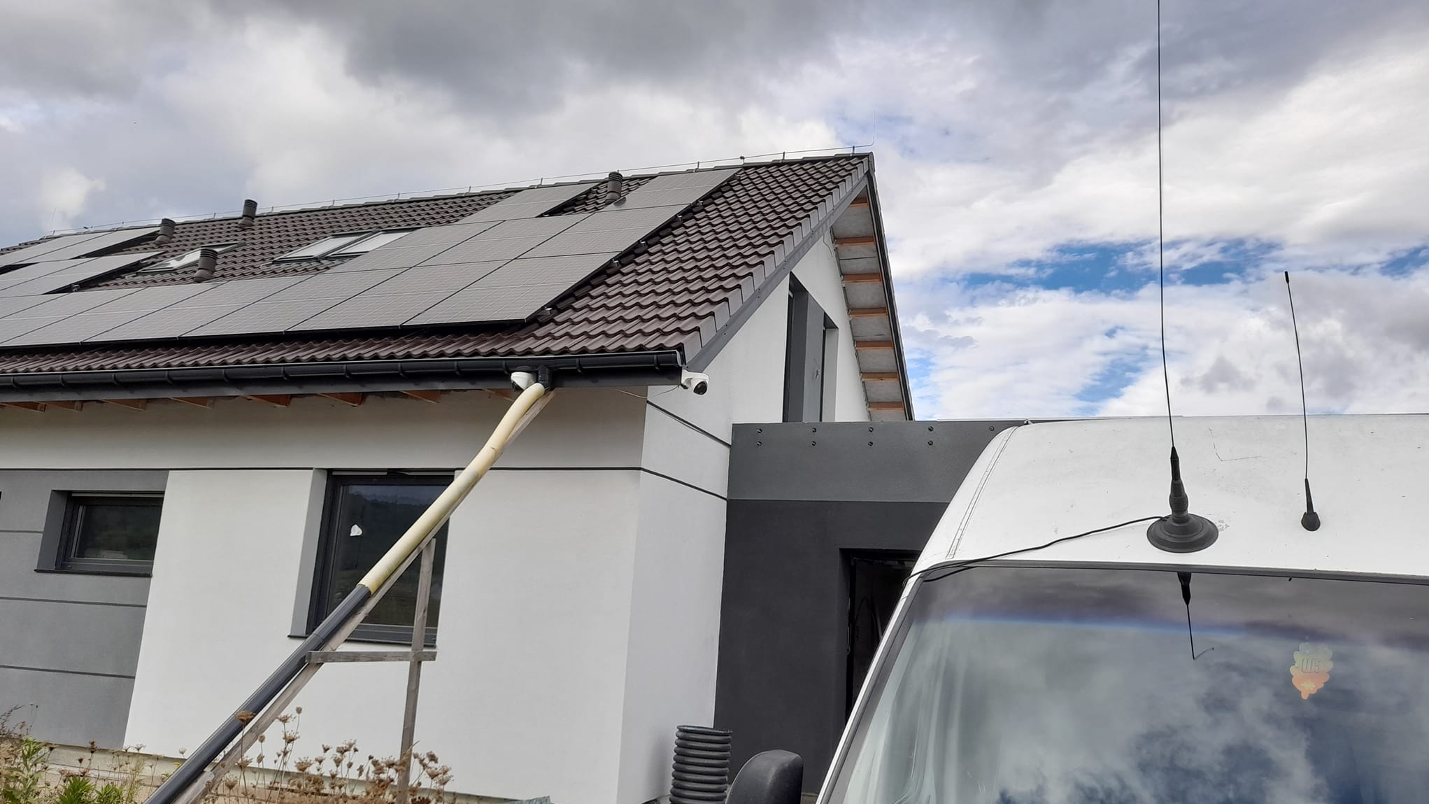 Dom jednorodzinny z panelami słonecznymi na brązowym dachu, drabina oparta o białą ścianę, widoczny fragment białego samochodu dostawczego z antenami na dachu.