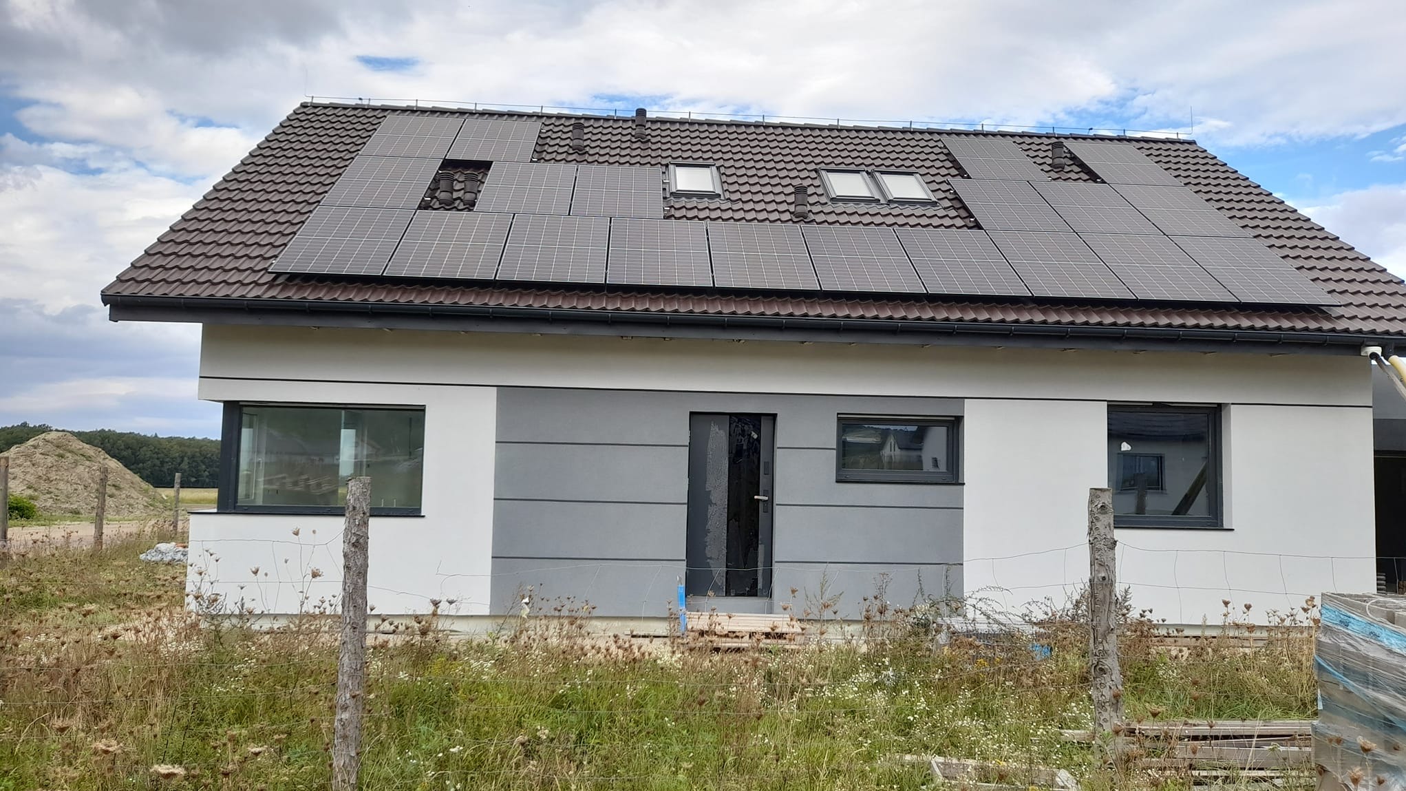 Nowoczesny dom jednorodzinny z brązową dachówką i panelami słonecznymi na dachu, widoczne okna dachowe, elewacja biało-szara, teren wokół domu częściowo zagospodarowany, w tle hałda ziemi.