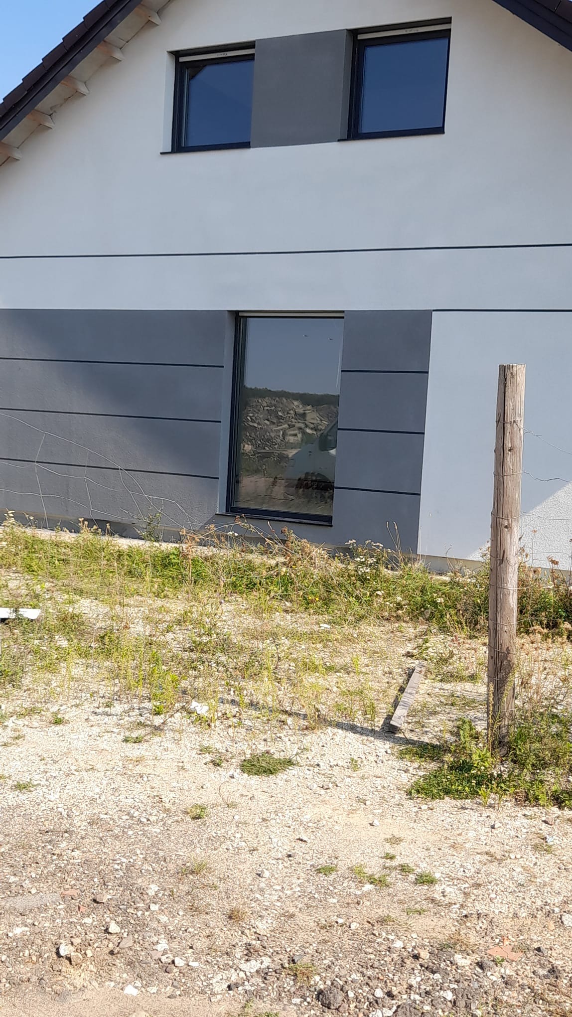 Dwukolorowa elewacja domu z widocznymi oknami, na pierwszym planie porośnięty teren i drewniany słupek ogrodzeniowy.