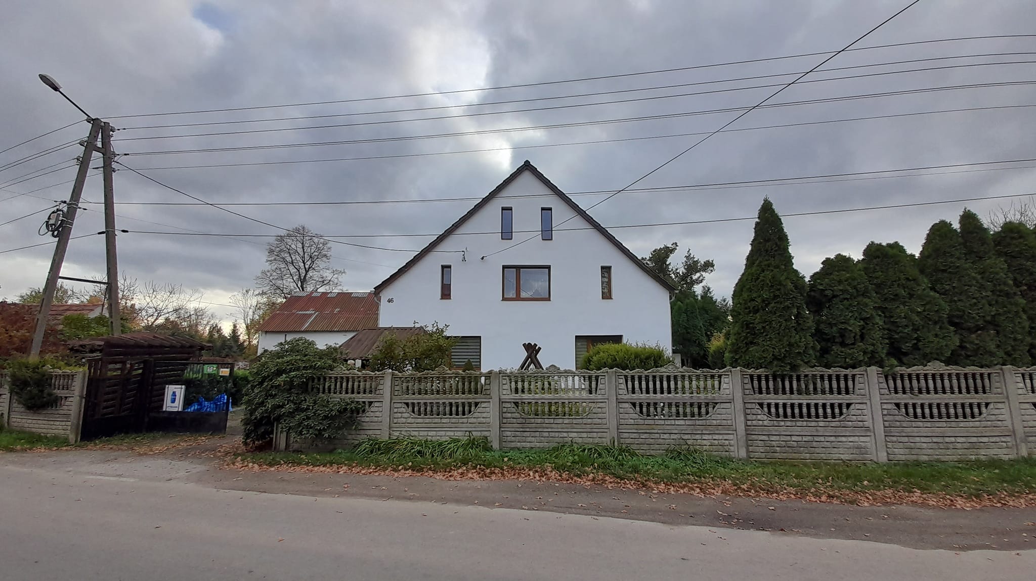 Biały, jednopiętrowy dom z szarym betonowym ogrodzeniem w pochmurny dzień, widoczne linie energetyczne i drzewa iglaste