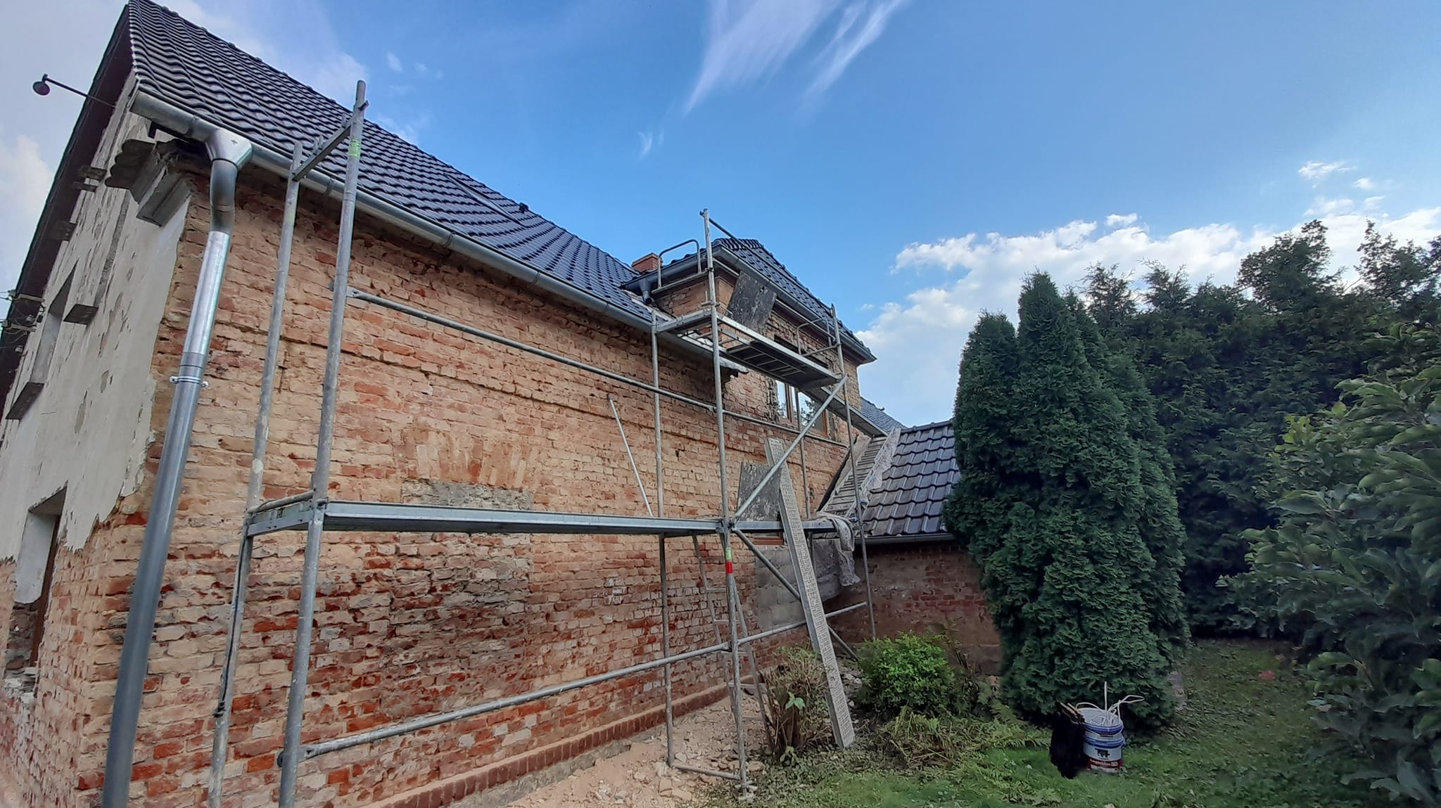 Dom z czerwonej cegły w trakcie prac remontowych na zewnątrz, widoczne rusztowanie, wiadro oraz fragmenty ocieplenia, nieotynkowane fragmenty elewacji.
