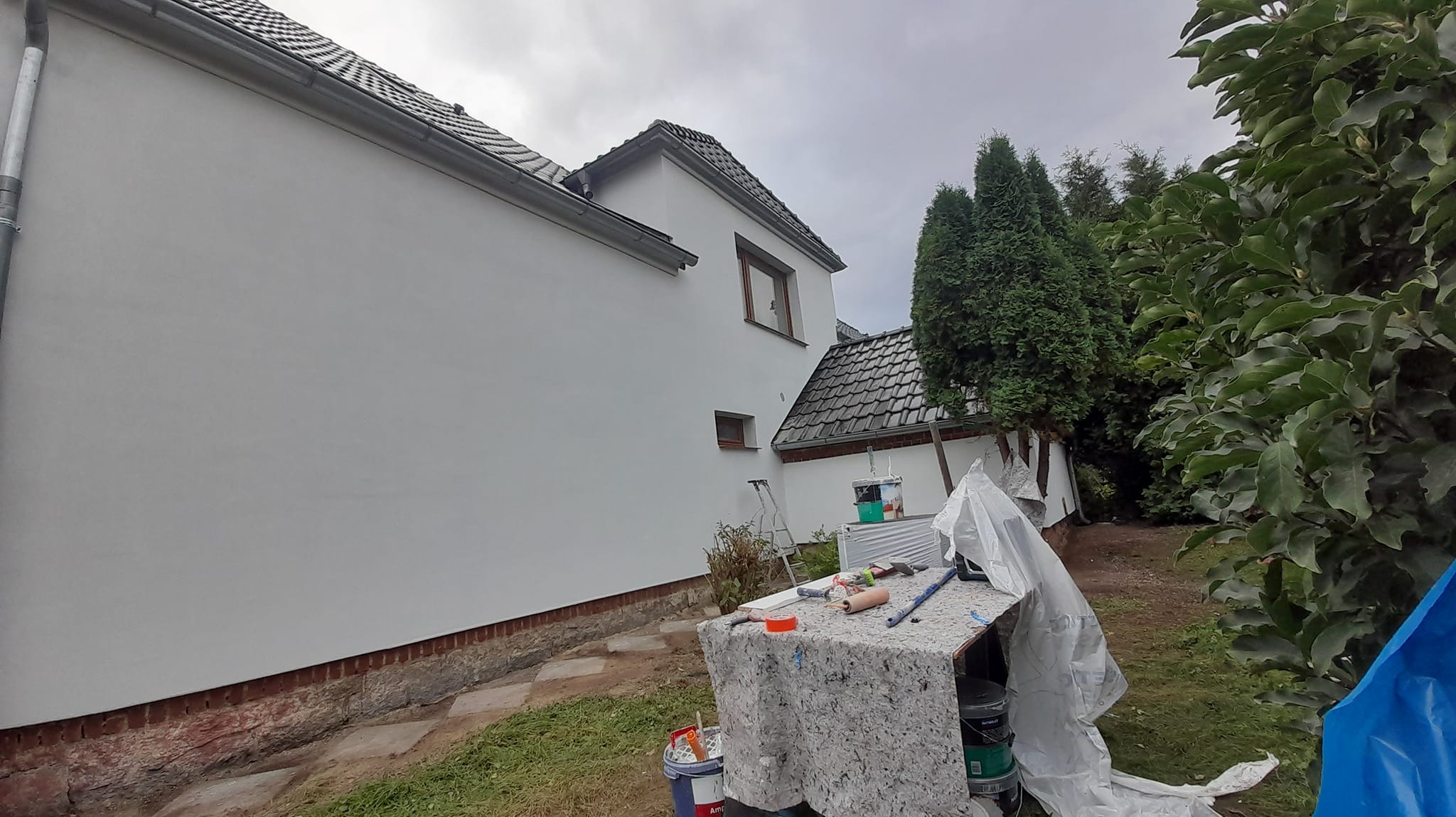 Świeżo pomalowana, biała ściana zewnętrzna domu jednorodzinnego z widocznymi narzędziami malarskimi, w tym wałkami i farbami, oraz drabina oparta o ścianę; w tle drzewa iglaste i pochmurne niebo.