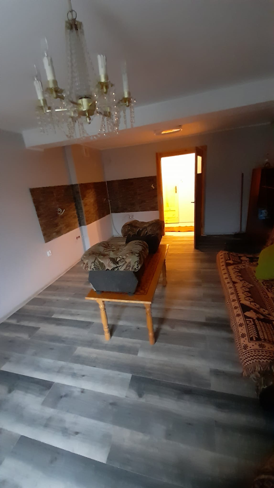 Wnętrze pokoju z panelami podłogowymi w odcieniach szarości, z widoczną ścianą z dekoracyjną okładziną imitującą cegłę, kryształowym żyrandolem i meblami w stylu retro.