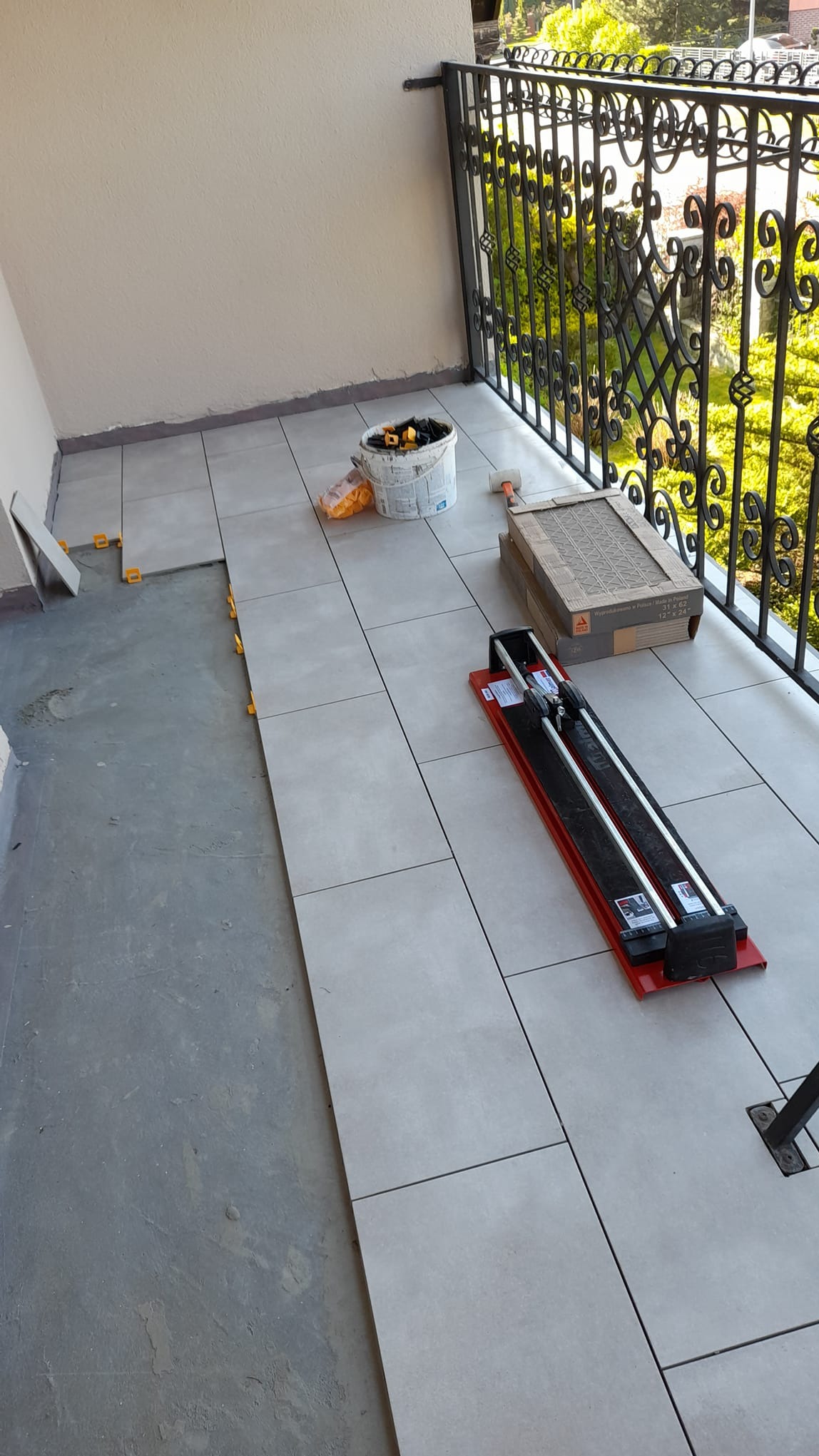 Balkon w trakcie kładzenia szarych płytek, widoczna przecinarka do płytek, wiadro z akcesoriami i paczka płytek.