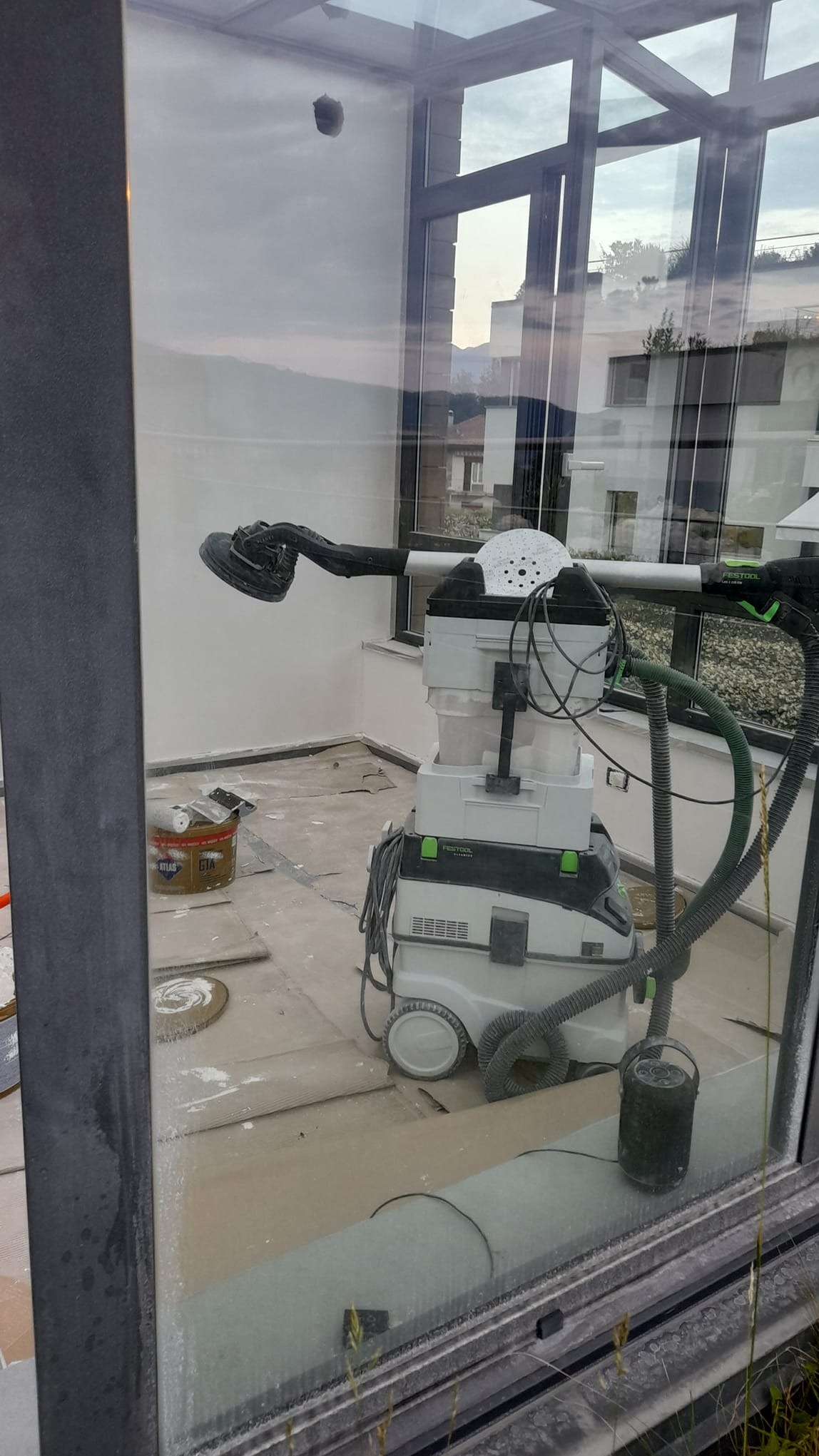 Szlifierka do gładzi z odkurzaczem przemysłowym Festool na podłodze w trakcie prac wykończeniowych na balkonie z widokiem na zabudowę mieszkalną.