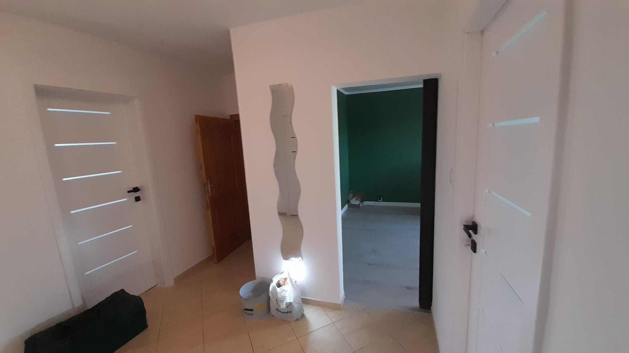 Wnętrze po remoncie: białe ściany, dwie białe drzwi z poziomymi przeszkleniami, brązowe drzwi, dekoracyjne lustro na ścianie, widoczne wiadro i worek z resztkami materiałów.