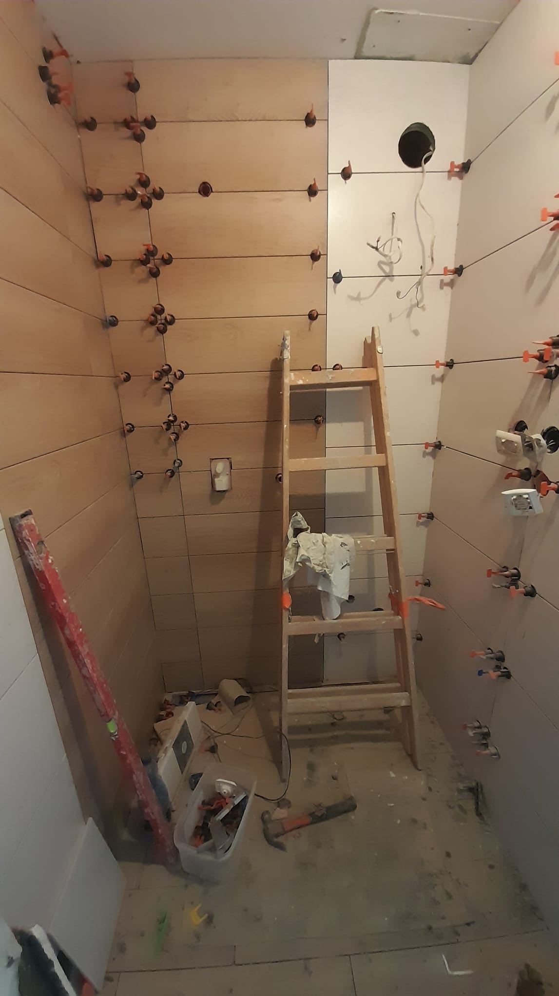 Remontowana łazienka z częściowo ułożonymi płytkami imitującymi drewno, widoczne poziomica, drabina i narzędzia glazurnicze z systemem poziomowania płytek.