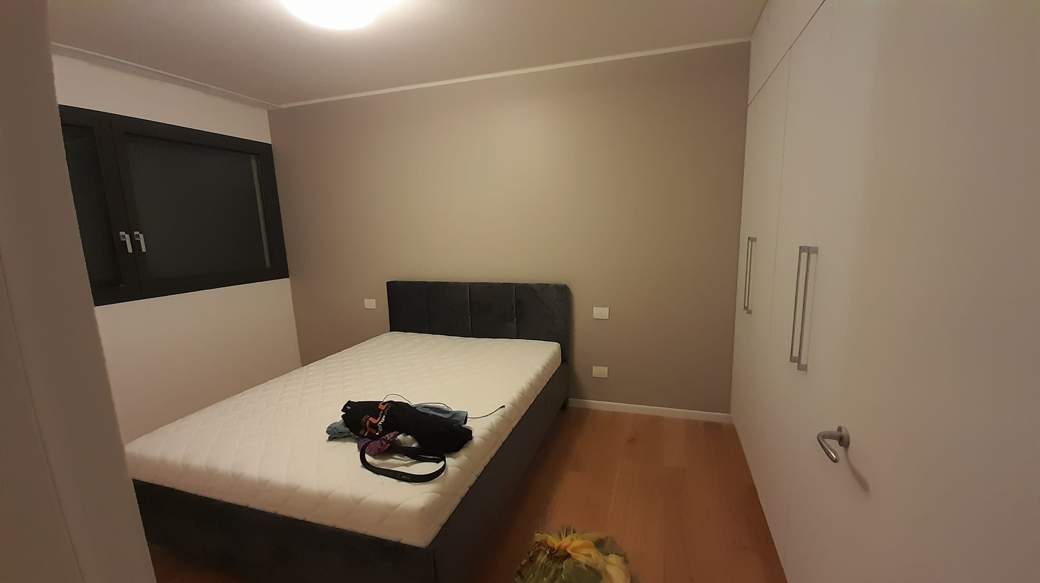 Wnętrze sypialni po malowaniu ścian na szaro, z zamontowanym oknem w czarnej ramie, nowym łóżkiem i białą szafą wnękową.