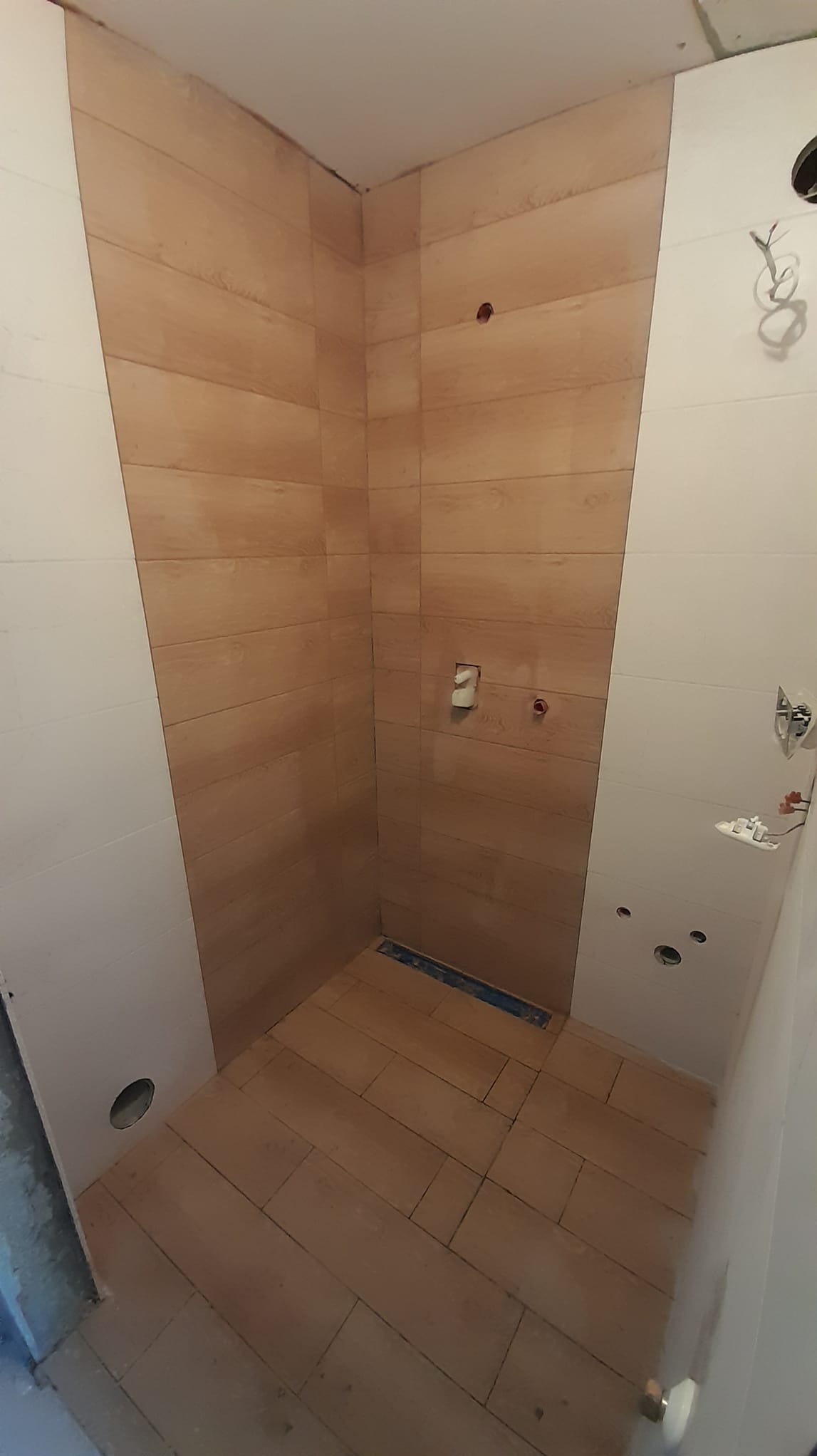 Wnętrze kabiny prysznicowej w trakcie wykańczania, z brązowymi płytkami imitującymi drewno na ścianach i podłodze, widoczne otwory na armaturę i odpływ liniowy, białe płytki na ścianach bocznych...