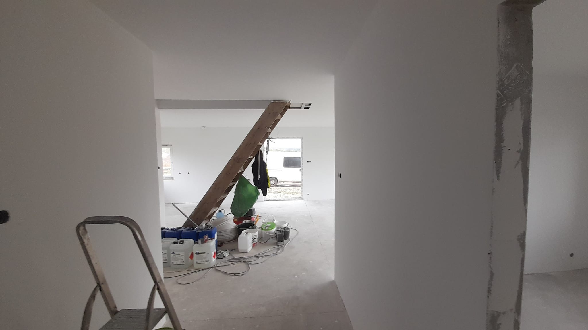 Wnętrze w trakcie remontu: świeżo pomalowane na biało ściany, drabina oparta o sufit, wiadro z farbą i akcesoria malarskie na podłodze.