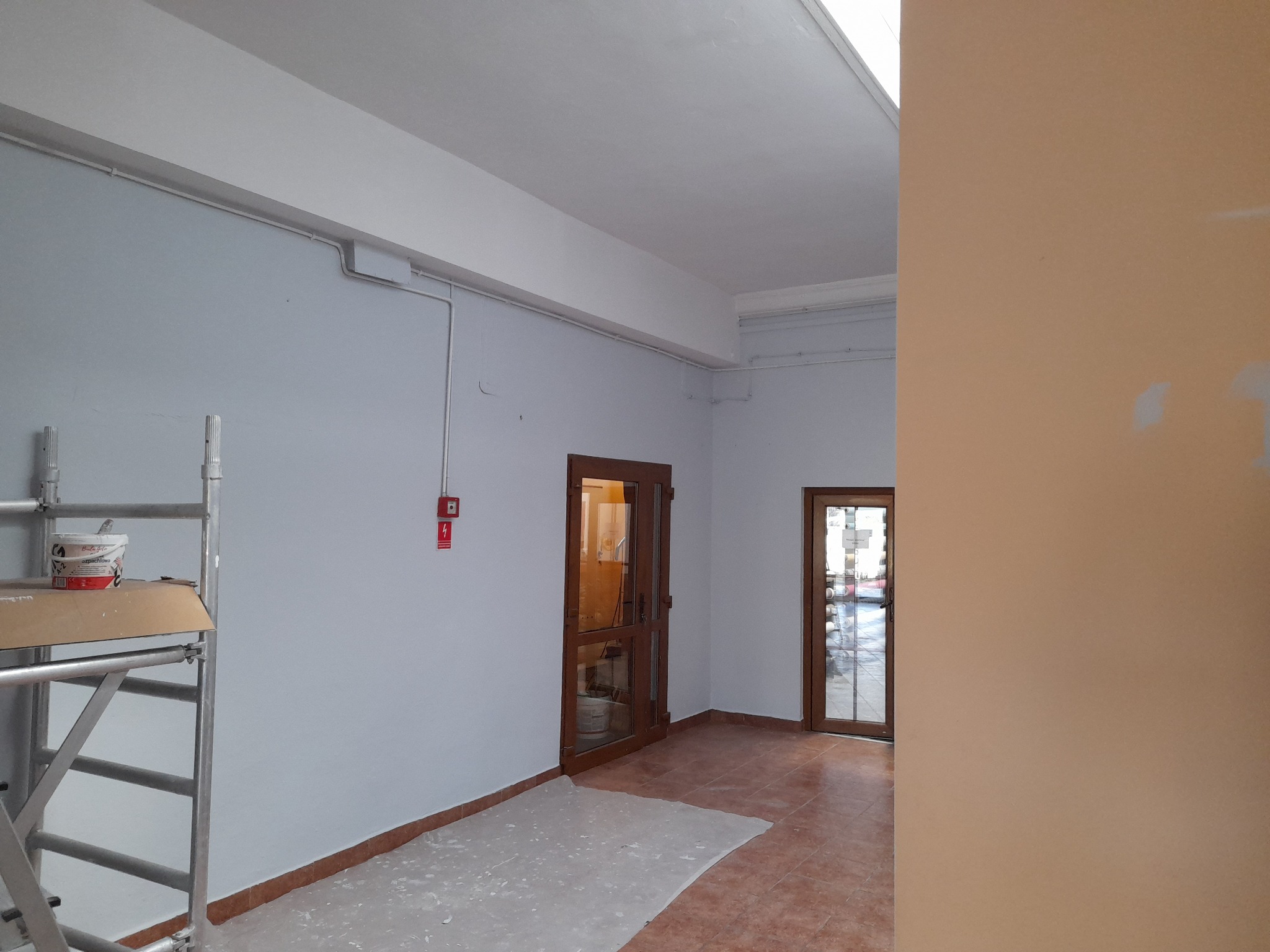 Wnętrze po świeżym pomalowaniu na jasnoszaro, widoczny aluminiowy ruszt z puszką farby, zabezpieczona folią podłoga i drewniane drzwi z szybami.