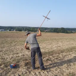 Pracownik w szarym ubraniu, kapeluszu i rękawicach wbija długi pręt z poprzeczką w ziemię na polu, obok leży niebieska skrzynka z narzędziami.