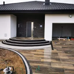 Nowoczesny dom z podjazdem wyłożonym drewnopodobną kostką, schody wejściowe z obrzeżami z czarnej kostki i białego grysu, widoczne narzędzia brukarskie i częściowo wykonane obrzeże rabaty.