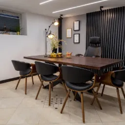 Nowoczesne biuro z ciemnym, drewnianym stołem konferencyjnym otoczonym czarnymi krzesłami, oświetlone lampami sufitowymi i lampą biurkową, z dekoracjami na stole i certyfikatami na ścianie.