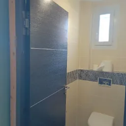 Nowoczesna łazienka z otwartymi, ciemnymi drzwiami z dekoracyjnymi liniami, jasnymi płytkami, mozaikowym pasem i wiszącą toaletą.