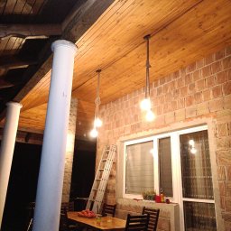 KONRAD POSŁUSZNY Instalacje Remonty i Wykończenia - Zadaszony taras z drewnianym sufitem, ceglanymi ścianami i kolumnami. Wiszące lampy na sznurze oświetlają drewniany stół z krzesłami. Opiera się o ścianę drabina. Na parapecie okna stoją doniczki...