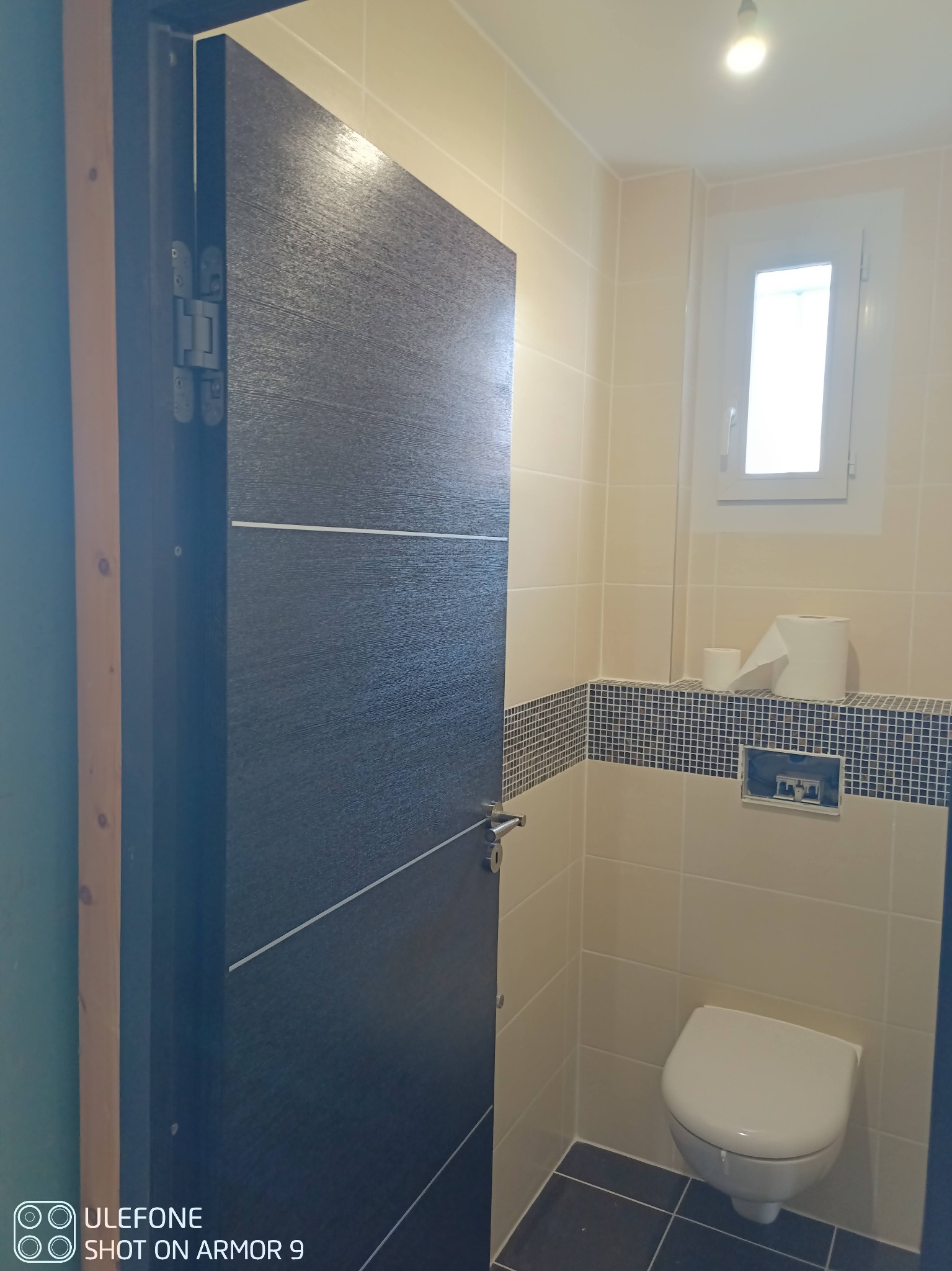 Nowoczesna łazienka z otwartymi, ciemnymi drzwiami z dekoracyjnymi liniami, jasnymi płytkami, mozaikowym pasem i wiszącą toaletą.