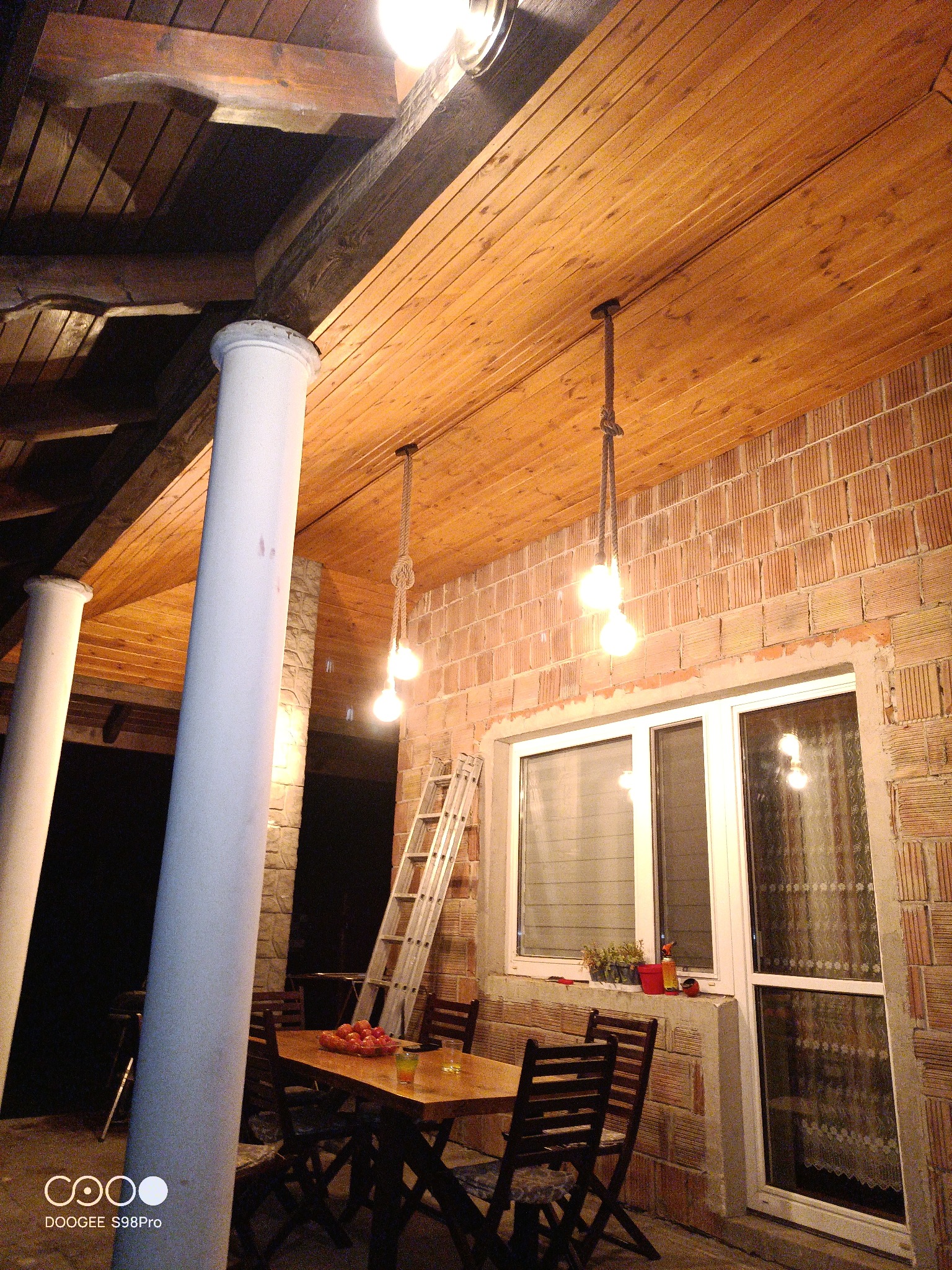 Zadaszony taras z drewnianym sufitem, ceglanymi ścianami i kolumnami. Wiszące lampy na sznurze oświetlają drewniany stół z krzesłami. Opiera się o ścianę drabina. Na parapecie okna stoją doniczki...