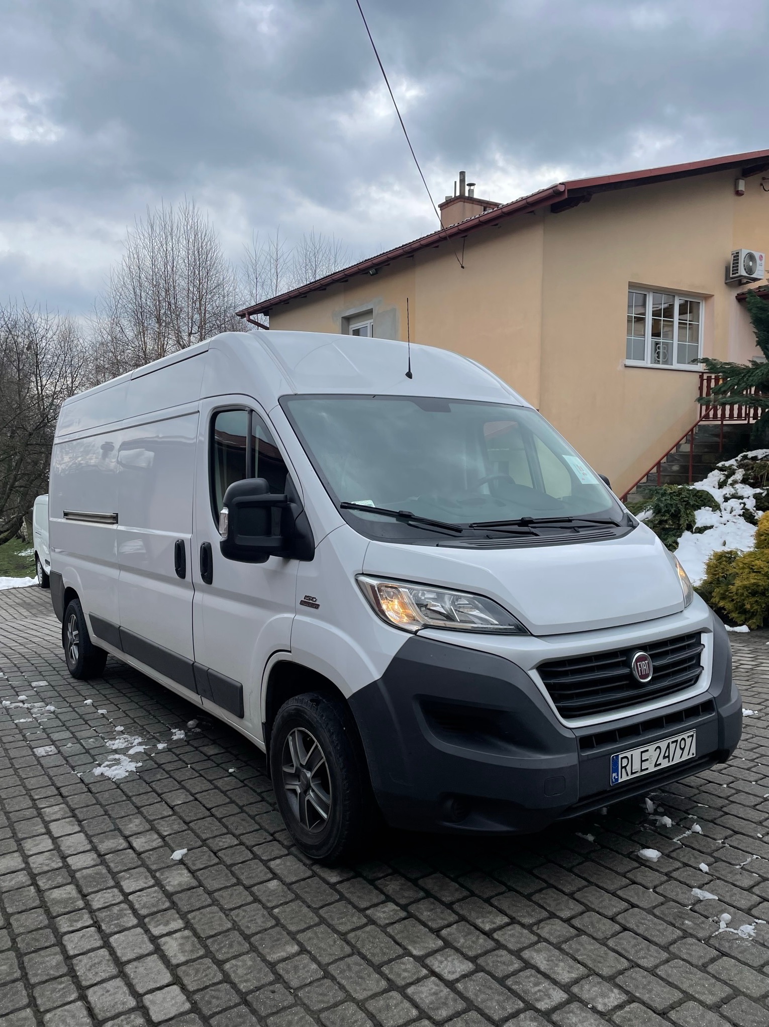 Biały samochód dostawczy Fiat Ducato zaparkowany na brukowanym podjeździe przed beżowym domem z czerwonym dachem w pochmurny dzień.